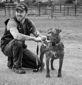 Bio black and white photo of Steffen Baldwin kneeling in a ranch setting with a dark-colored dog